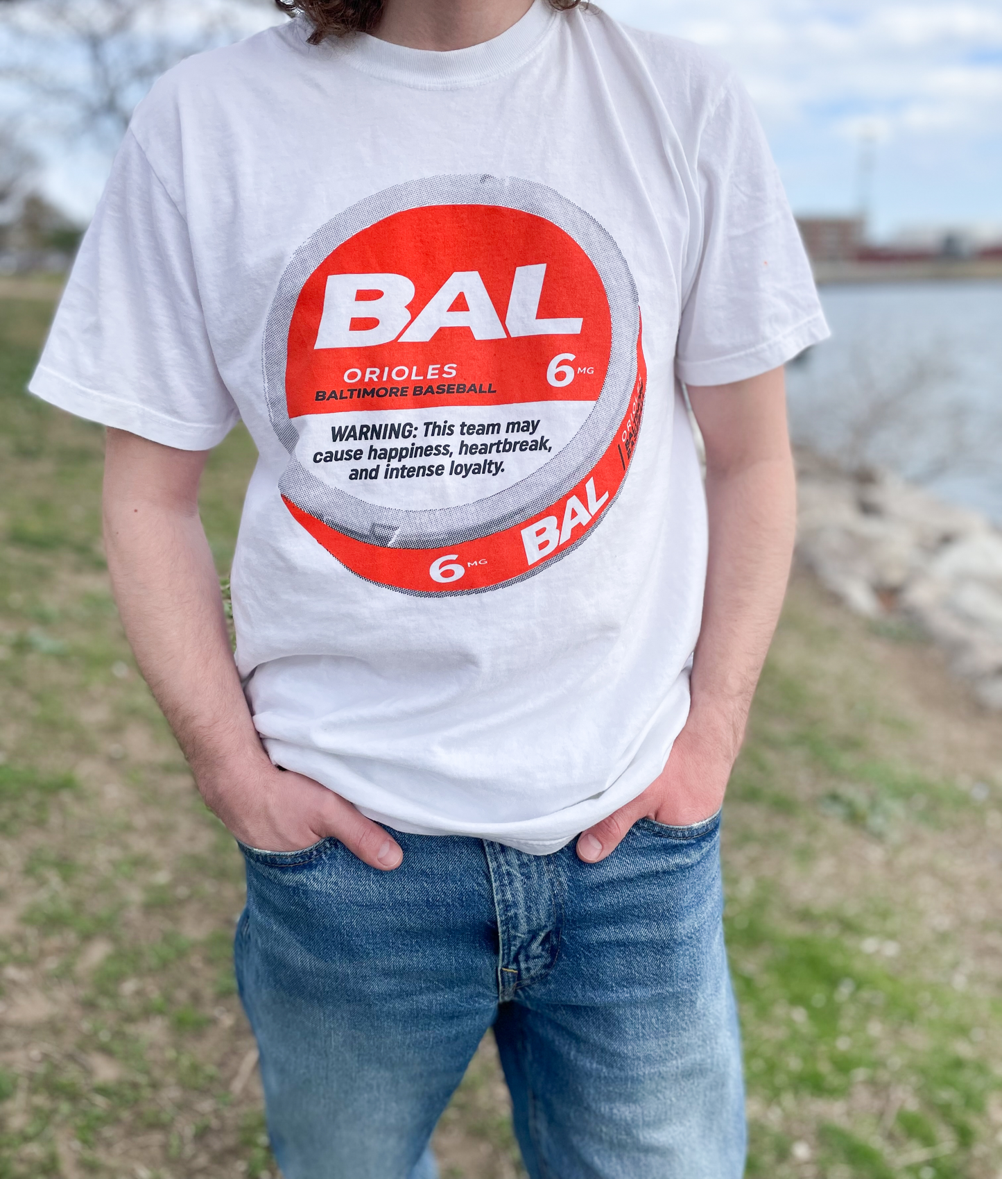 person wearing a white t shirt and blue jeans on a grassy knoll. the white t shirt is featured with an orange disc and text that is black.