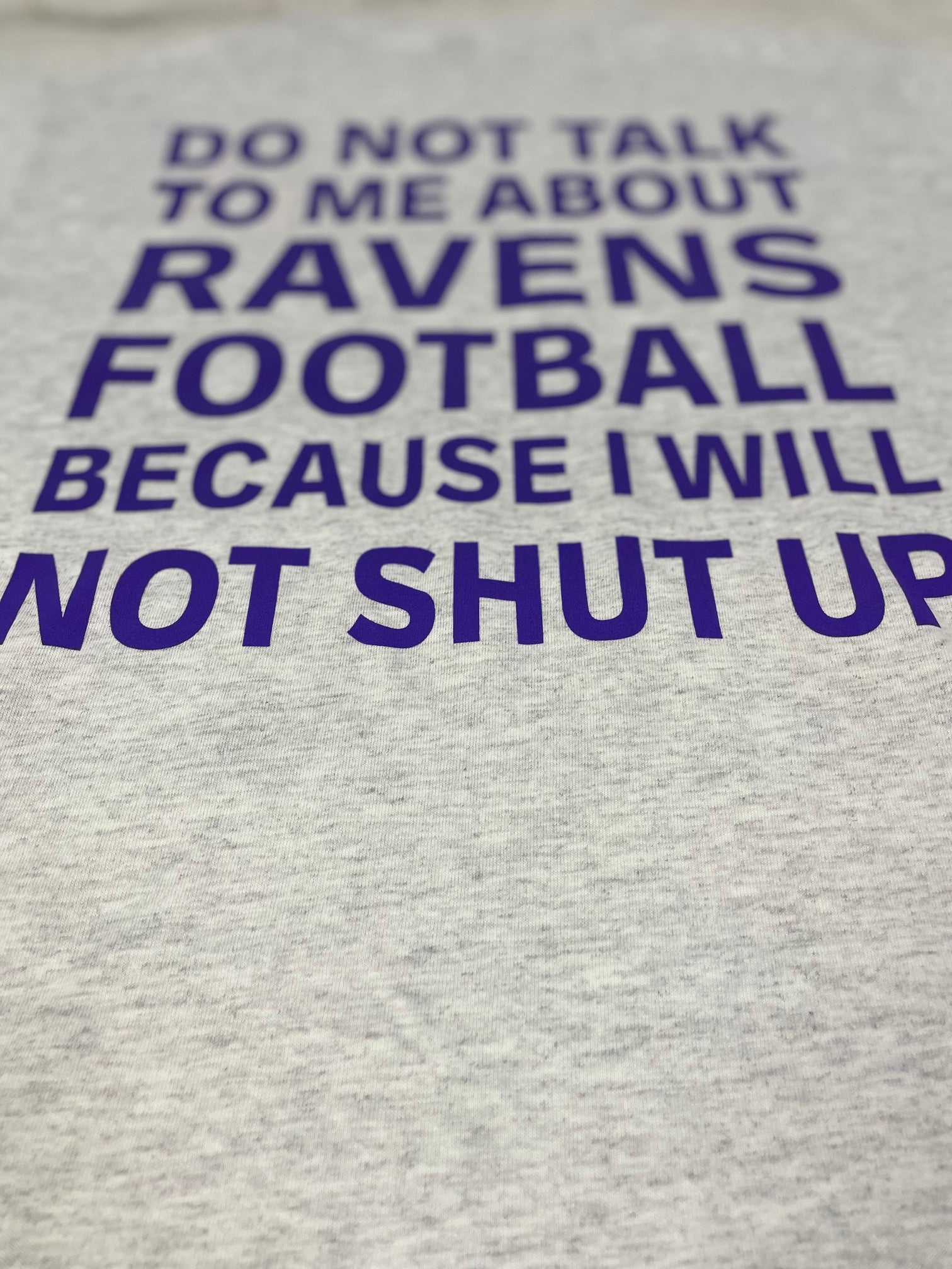 A close up of T-shirt that is Ash White in color, showing the back of the t-shirt with purple text that says "DO NOT TALK TO ME ABOUT RAVNES FOOTBALL BECAUSE I WILL NOT SHUT UP"